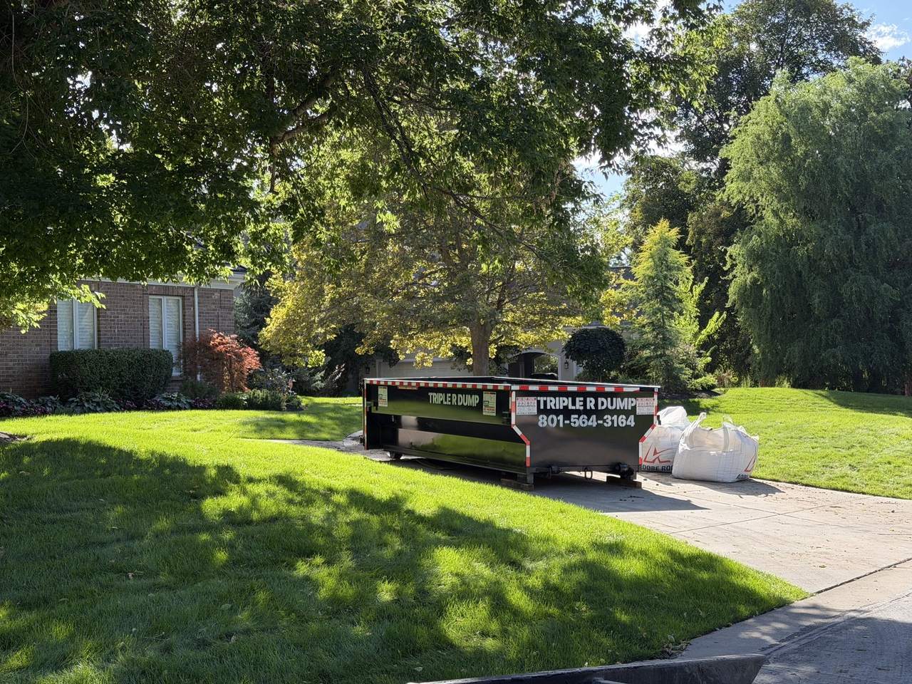 Triple R Dump roll-off dumpster on a driveway beside a large lawn and bulk bags in Cottonwood Heights, Utah