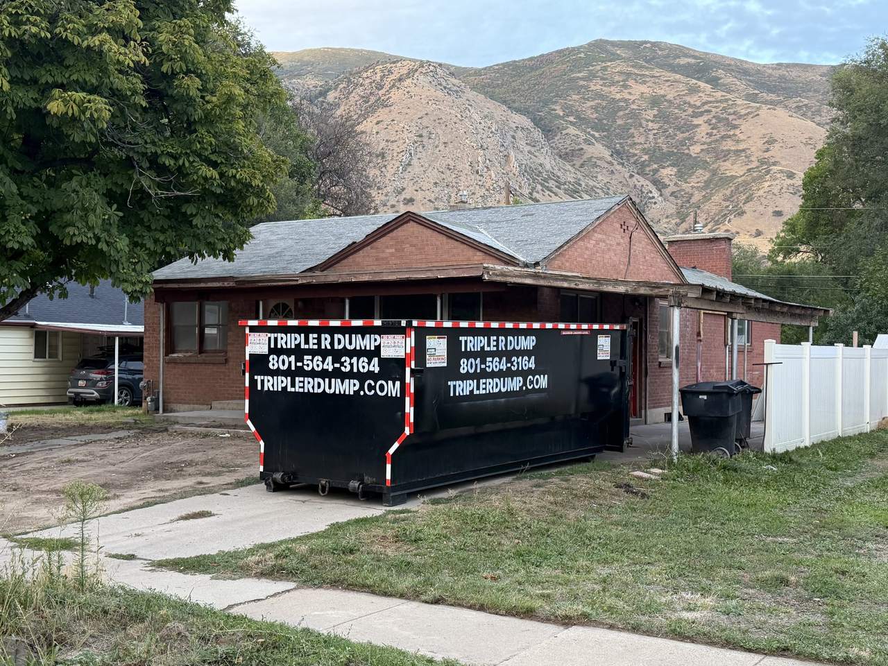 Triple R Dump roll-off dumpster in front of a brick home in Springville, Utah, with the mountains behind